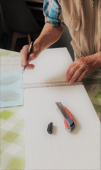 Fotografia 7 przedstawia kadr z zajęć arteterapeutycznych - klient odmierza linijką na białym styropianie szerokość elementu. Na styropianie leży nożyk introligatorski w stalowo - pomarańczowym kolorze