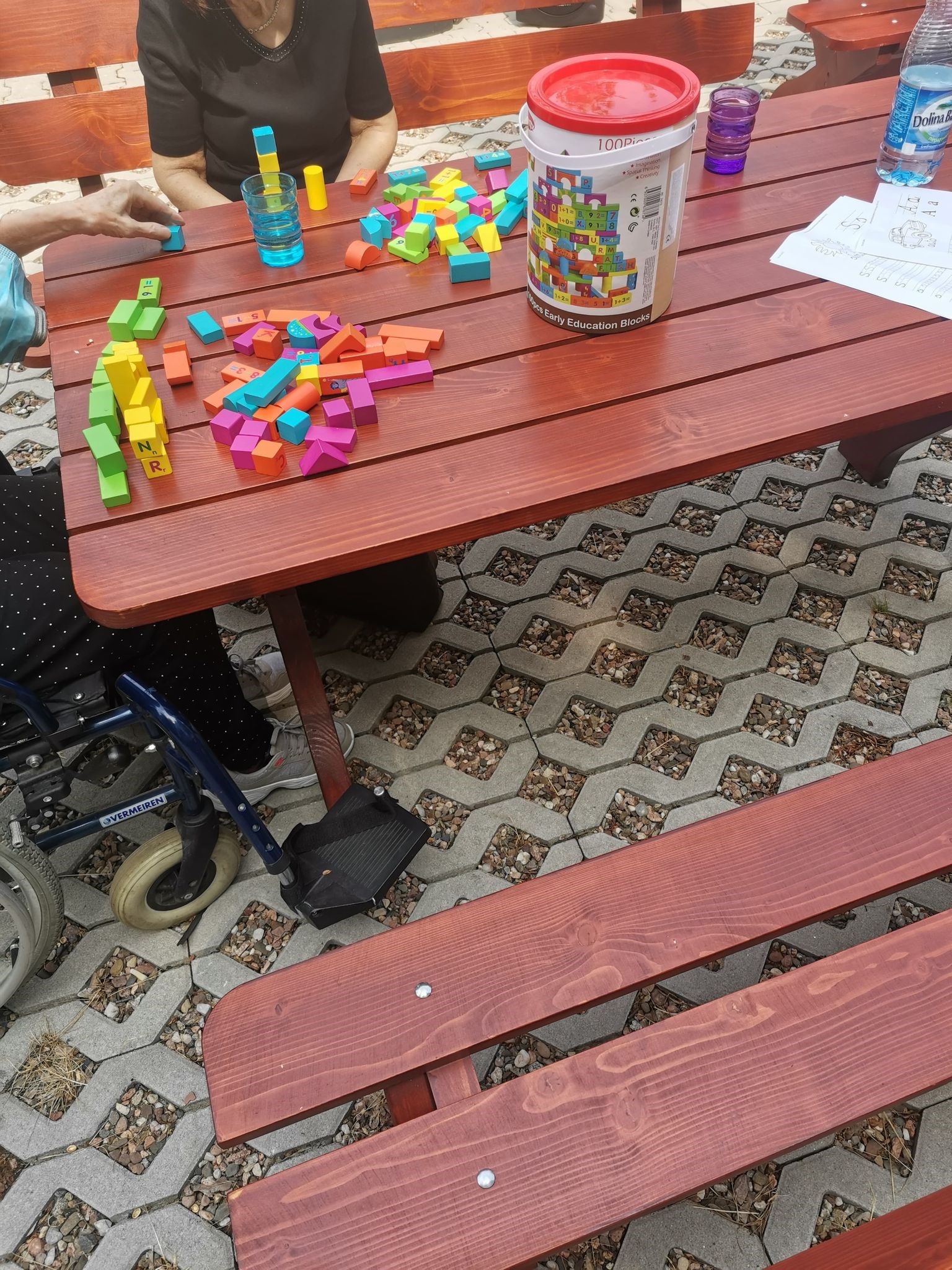  Kadr z zajęć trenujących koordynację wzrokowo – ruchową. Zdjęcie brązowego, drewnianego stołu, na którym znajdują się klocki w kolorze fioletowym, żółty, zielonym, pomarańczowym i niebieskim.