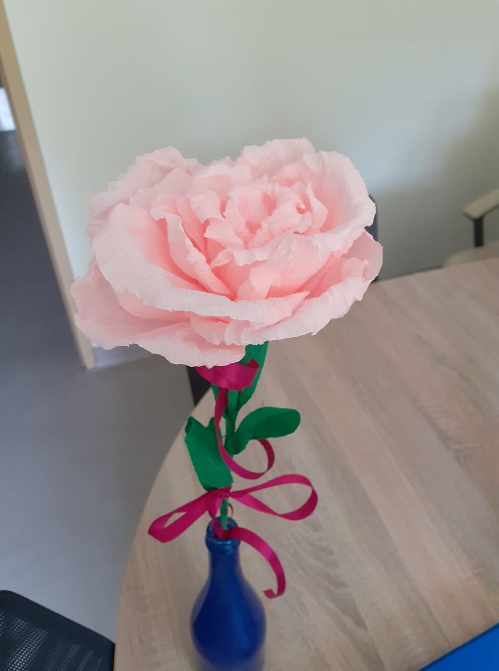 Obraz przedstawia stojący w niebieskim wazoniku, ręcznie zrobiony z bibuły kwiat (różową różę przewiązaną wstążką) wykonany podczas zajęć dla jednej z kliente DDP z okazji jej imienin.
