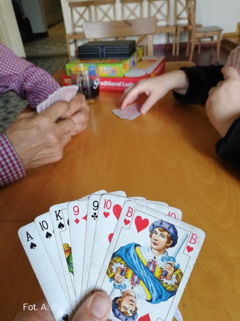 Na zdjęciu na pierwszym planie talia 7 kart rozłożona w wachlarz, na drugim planie stół z innymi grami planszowymi po obu stronach siedzą osoby grające w grę tysiąc