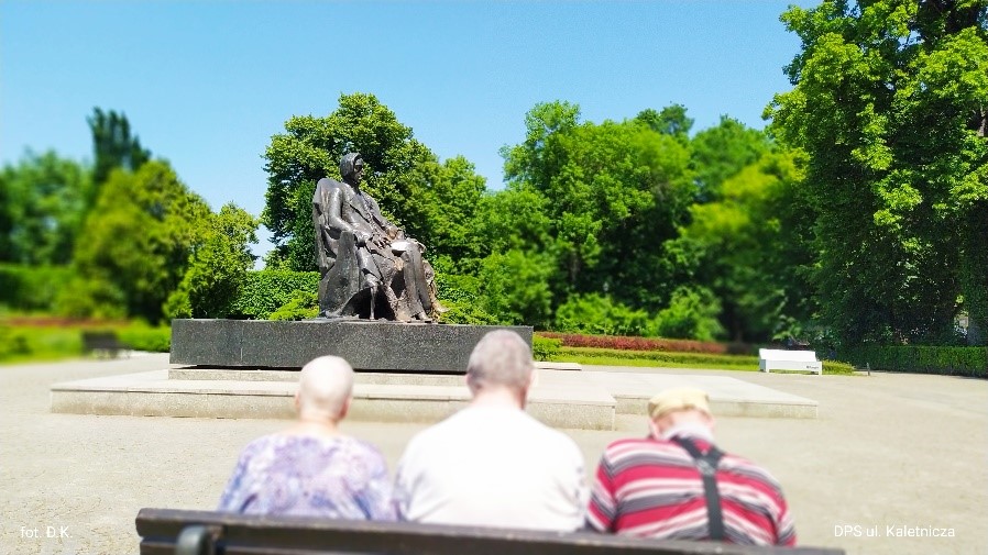 Na zdjęciu, na pierwszym planie 3 sylwetki osób siedzących na ławce, widzianych z tyłu. Przed nimi plac z pomnikiem Fryderyka Chopina. W tle zielone drzewa