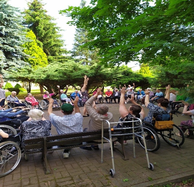 Grupa mieszkańców siedzącą na ławkach w ogrodzie i podnosząca ręce do góry