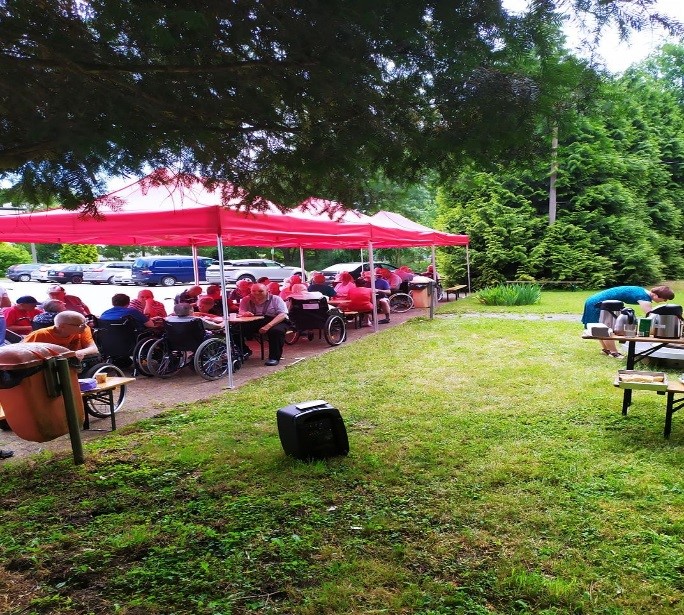 Grupa mieszkańców DPS siedzących przy stołach w ogrodzie i jedzących grillowane przysmaki