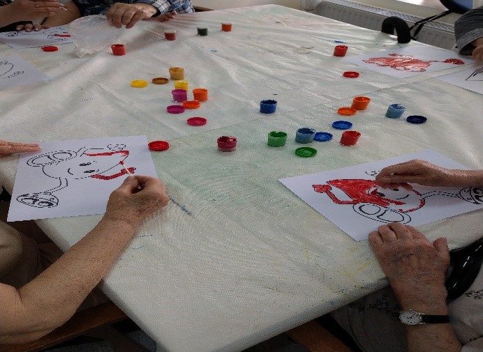 Fotografia przedstawia stół pokryty białą ceratą na którym widać ręce seniorów trzymające pędzle podczas malowania kartek z rysunkiem kropli krwi. Na stole widać porozkładane małe pojemniki z różnokolorowymi farbami