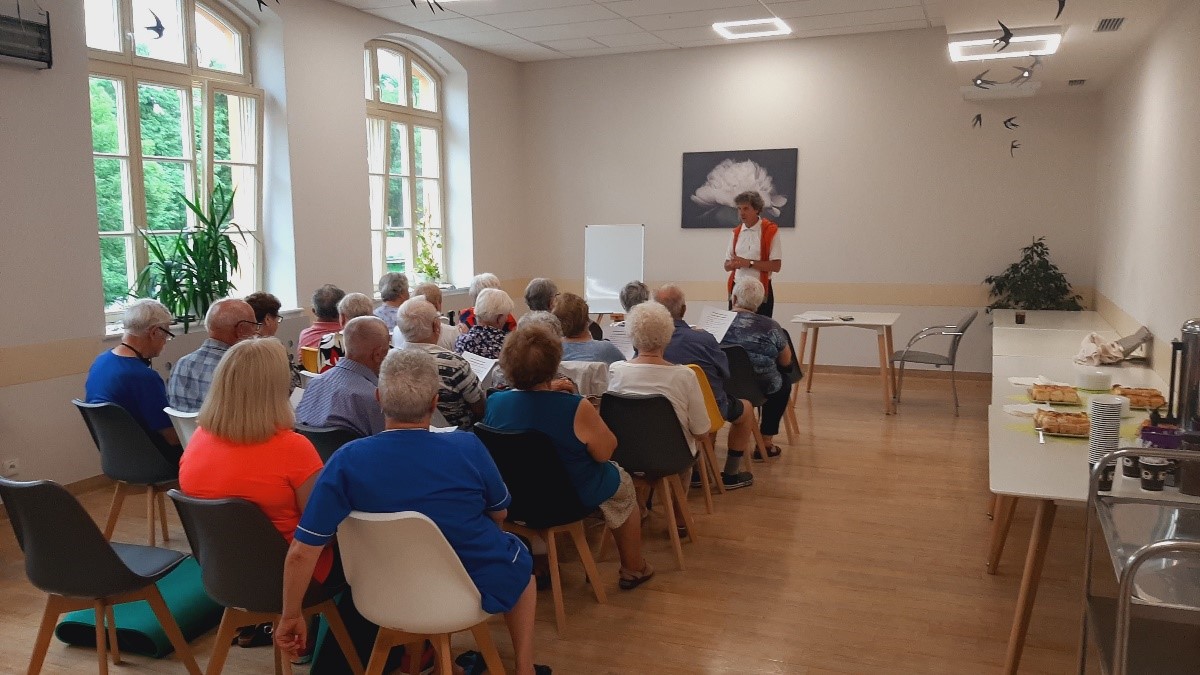 Sala wykładowa w kolorze białym. Na pierwszym tle seniorzy siedzący na plastikowych krzesełkach w kolorze szarym i białym, w drugim tle osoba prowadząca wykład. Po prawej stronie krzesła stoły na których znajduje się słodki poczęstunek dla gości oraz naszych seniorów