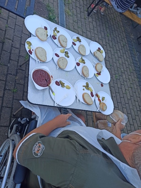 Na zdjęciu widać tacki z pieczywem oraz sosami, które leżą na stole. Przy stoliku siedzi jeden z mieszkańców.