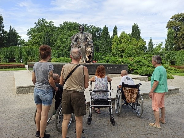 Zdjęcie przedstawia mieszkańców dps, którzy podziwiaja pomnik fryderyka Chopina w otoczeniu roślinności.