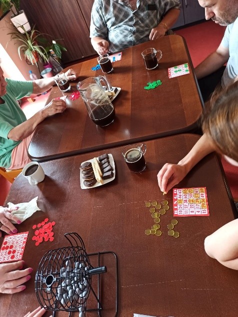 Na zdjęciu widzimy mieszkańców grających w grę BINGO. Na brązowym stole leżą: kawa, ciasteczka na białym talerzu oraz karty i żetony.