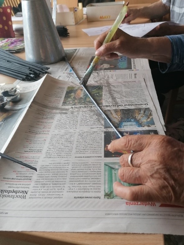 Mieszkanka siedzi w pracowni terapii zajęciowej i maluje srebną farbą rurki wykonane z gazety