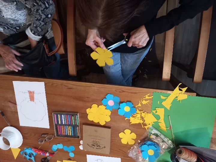 fotografia przedstawia stół, na którym widać ręce seniorów przy wycinaniu papierowych kwiatków. Na stole leżą już wycięte kolorowe papierowe kwiatki i kredki