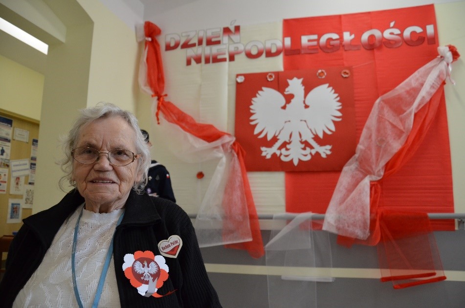Zdjęcie 2.Na pierwszym planie podopieczna DDP z przypiętym kotylionem, a w tle Godło Polski z biało-czerwoną flagą umieszczone na ścianie. 