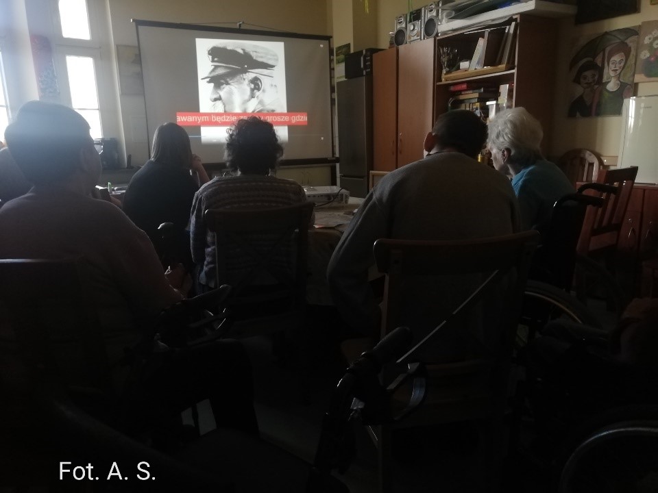 Na zdjęciu kilku mieszkańców siedzący w pokoju terapii zajęciowej, patrzących na ekran do projektora w tle wizerunek Józefa Piłsudskiego