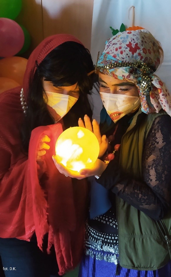 Na zdjęciu dwie kobiety ubrane w kolorowe stroje trzymające w rękach świecącą na żółto kulą. W tle różnokolorowe balony