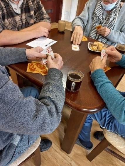Zdjęcie nr 4. Przedstawia mieszkańców grających w karty na brązowym stoliku. Leży na nim również poczęstunek