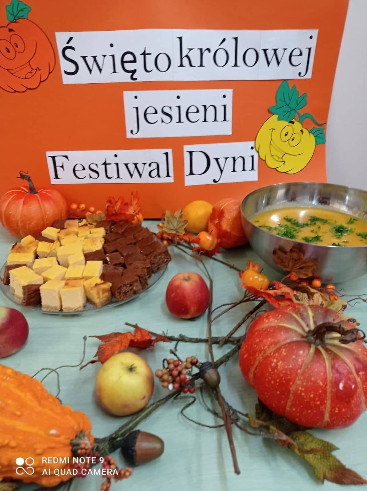 na pierwszym planie , dekoracja z dyni, jabłek i gałązek, miska z zupą dyniową oraz patera z ciastem . W tle na pomarańczowym kartonie napis: święto królowej jesieni – festiwal dyni