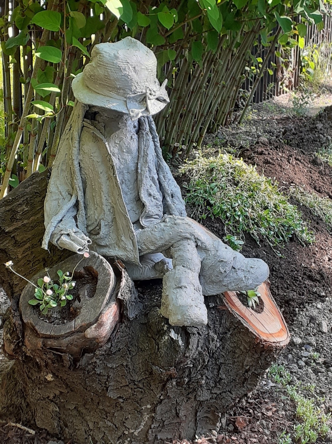 Obraz 1. Przedstawia szarą rzeźbę pt.: „Włóczykijka” wykonaną na zajęciach terapeutycznych z cementu. Rzeżba umieszczona jest na dniu drzewa