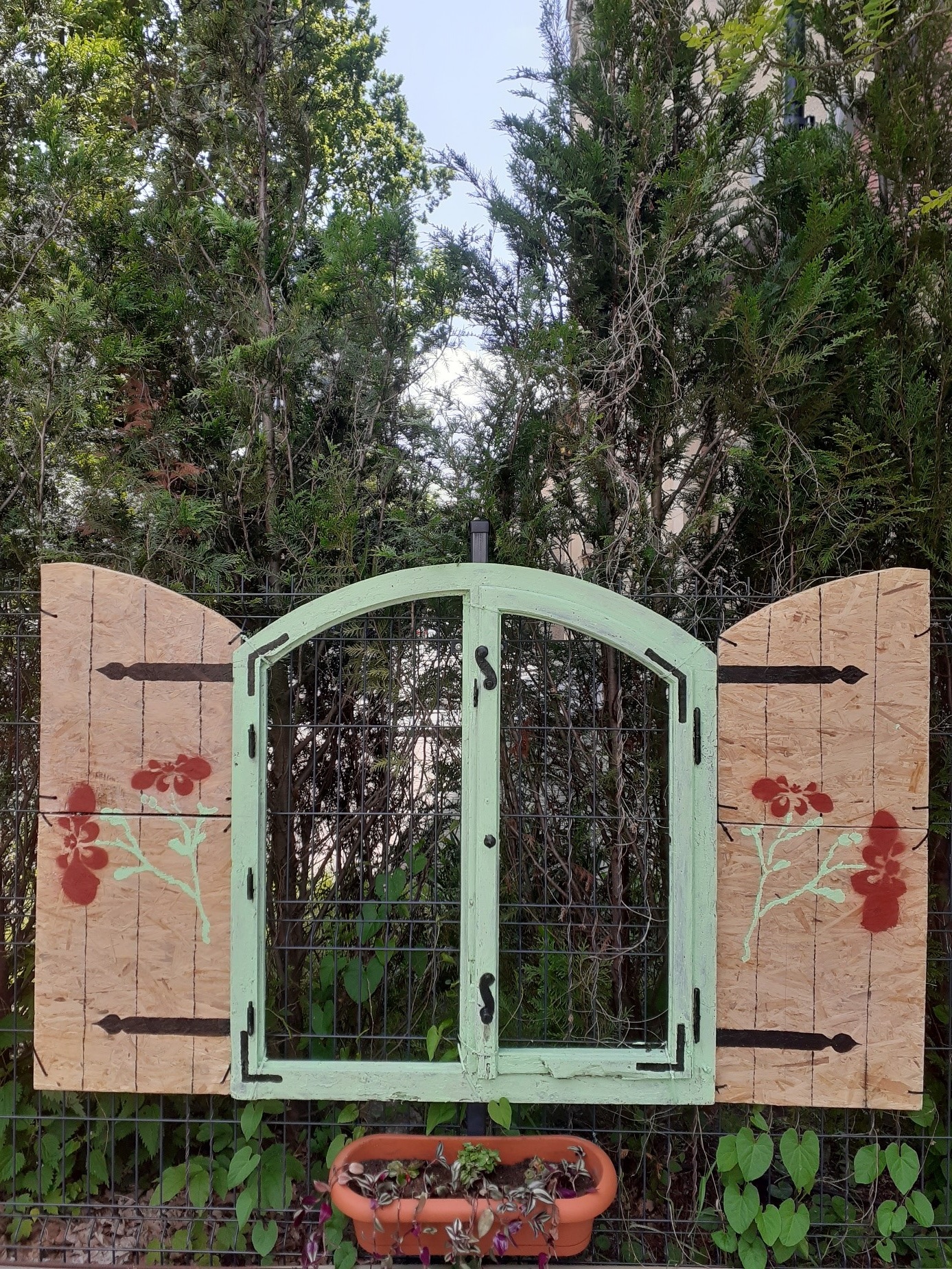 Obraz Przedstawia „Tajemnicze okno”  w ogrodzie DDP. Okno z zielonymi ramami i brązowymi okiennicami na tle drzew.