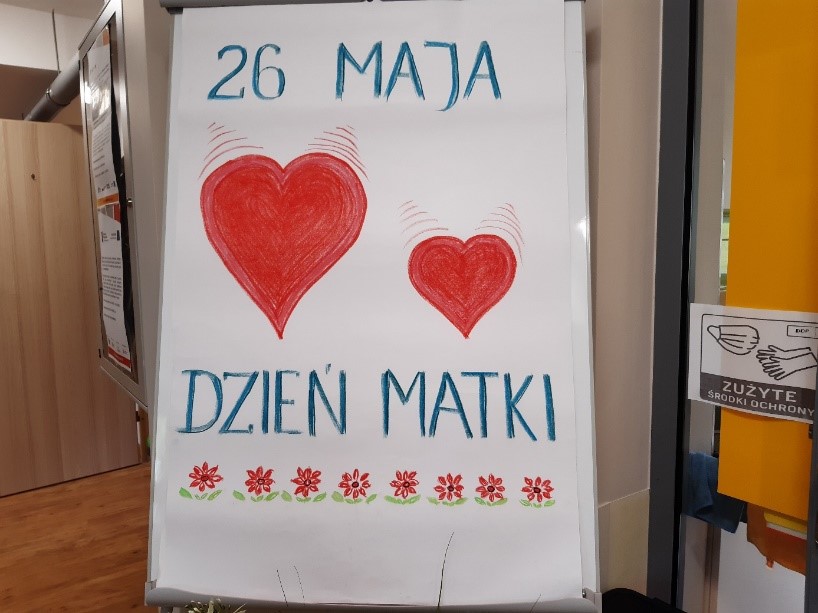 Plakat . Na dużej białej kartce napis 26 maja Dzień Matki oraz dwa czerwone serca o różnej wielkości. Dodatkowo dół plakatu udekorowany ośmioma czerwonymi kwiatuszkami