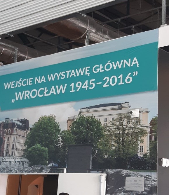 Wystawa pod tytułem „Wrocław 1945-2016”. Plakat z napisem "Wejście na wystawę główną. Wrocław 1945-2016”. Poniżej zdjęcie jednej z ulic Wrocławia -budynki, drzewa