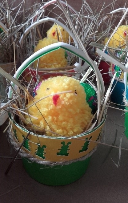 Fotografia przedstawia na pierwszym planie zielono-beżowe koszyczek z żółtym kurczakiem z włóczki, w tle żółte kurczaczki w koszykach