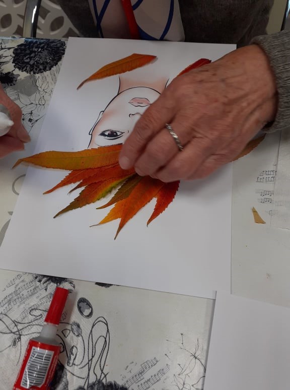  Zdjęcie 1 Fryzura z jesiennych liści. Na zdjęciu widzimy wydrukowaną twarz kobiety. klientka przykleja liście tworząc w ten sposób włosy, fryzurę. Używa do tego kolorowych liści w barwach żółto- czerwono- pomarańczowych. Obraz jest na białym tle.