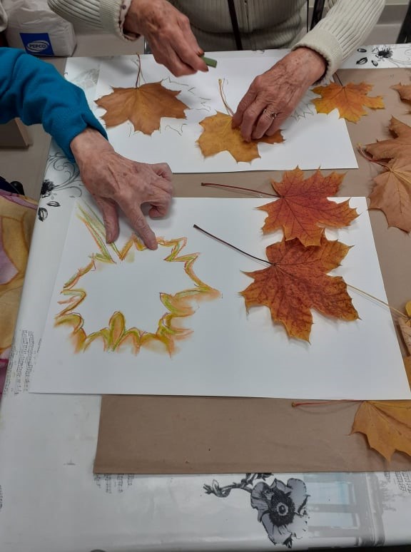Zdjęcie 2 Liście malowane pastelami suchymi. Na zdjęciu widzimy jak jedna z klientek odrysowuje kształt liścia. Druga klientka palcem rozmazuje kolorowe kształty liścia. Po prawej stronie zdjęcia widzimy dwa żółto- czerwone liście klonu.