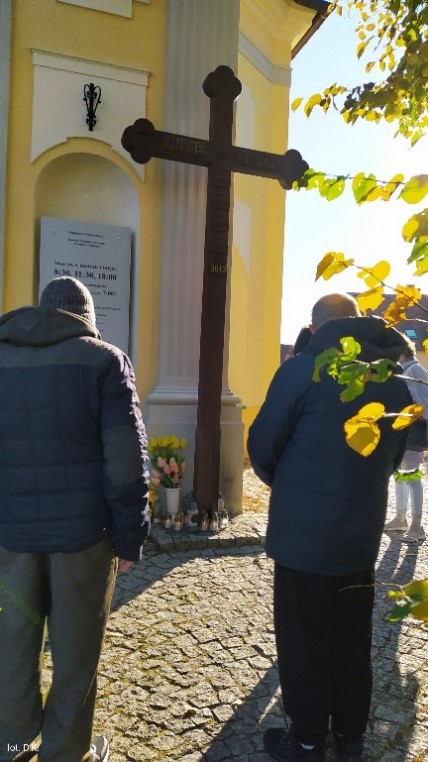 Na zdjęciu kilka osób stojących przy wejściu do kościoła, w tle duży drewniany krzyż