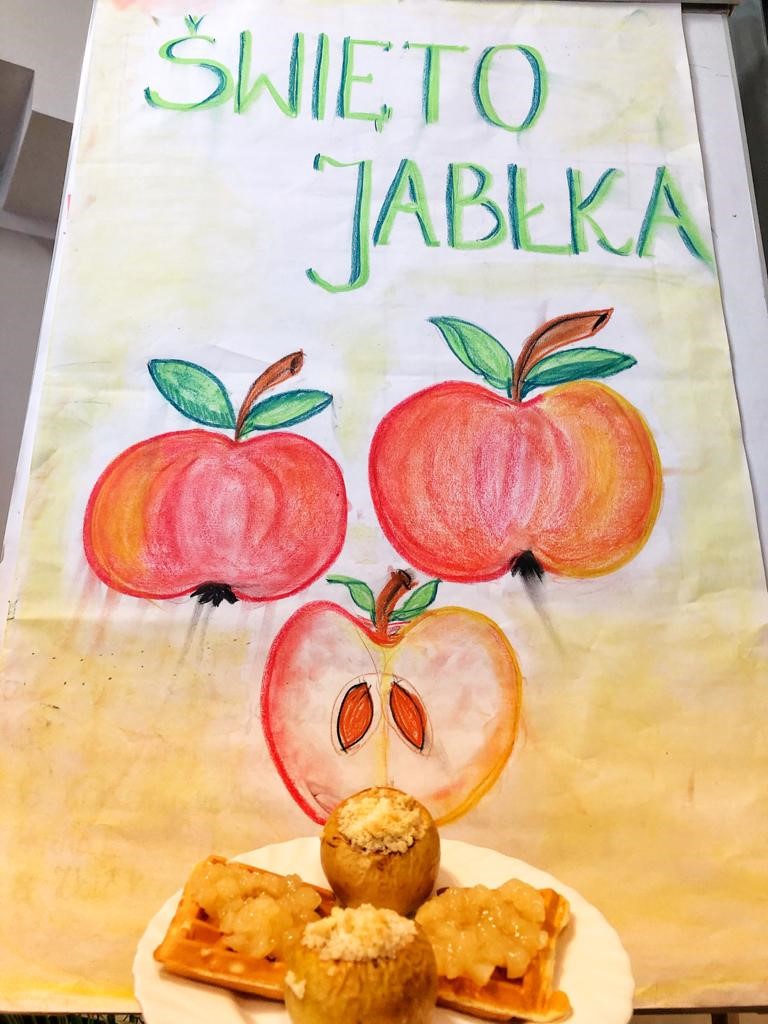 Ręcznie robiony plakat z okazji Święta Jabłka. Pod plakatem talerz, na którym znajdują się dwa pieczone jabłka z kruszonką oraz dwa gofry polane musem z jabłek.