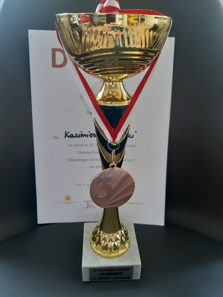  Nagrody zdobyte przez naszego klienta podczas Turnieju Gry w Tysiąca organizowanego przez DPS przy ul. Rędzińskiej. Na pierwszym planie złoty puchar i zawieszony na nim brązowy medal za zdobycie 3. miejsca. Na dalszym planie pamiątkowy dyplom.
