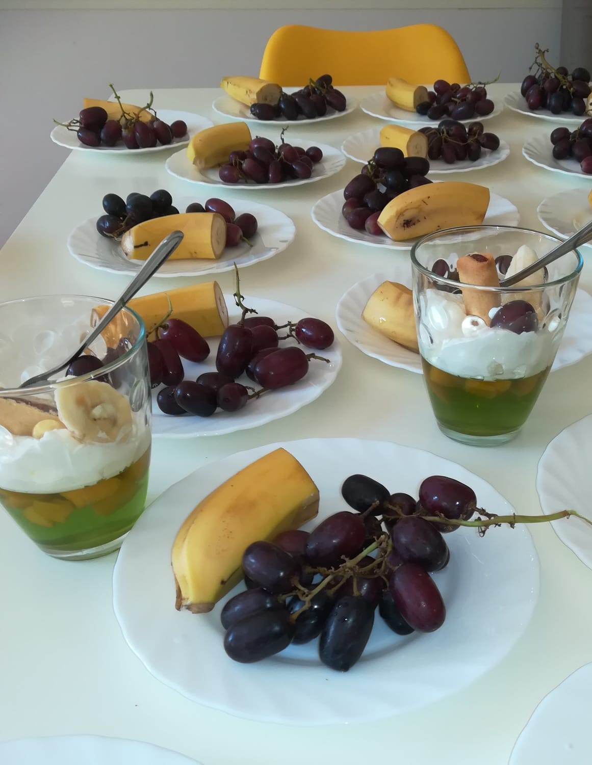 Na białym stole leżą talerzyki z owocami (banany i winogrona) oraz pucharki z deserem na bazie galaretki z owocami, ozdobionej bitą śmietaną i świeżymi owocami.
