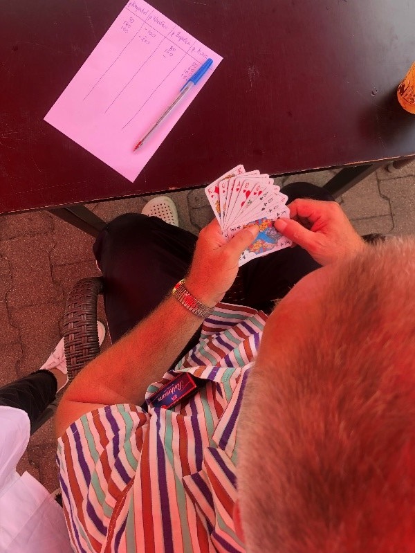 Na zdjęciu mieszkaniec siedzący na krześle przy stole,  trzyma rozłożoną talię kart. Na brązowym stole, leży biała kartka z zapisanymi wynikami oraz długopis z niebieską zakrętką