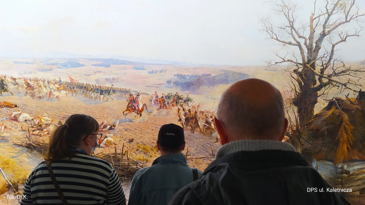 Na zdjęciu na pierwszym planie sylwetki trojga mieszkańców, na drugim planie obraz „Bitwa pod Racławicami”