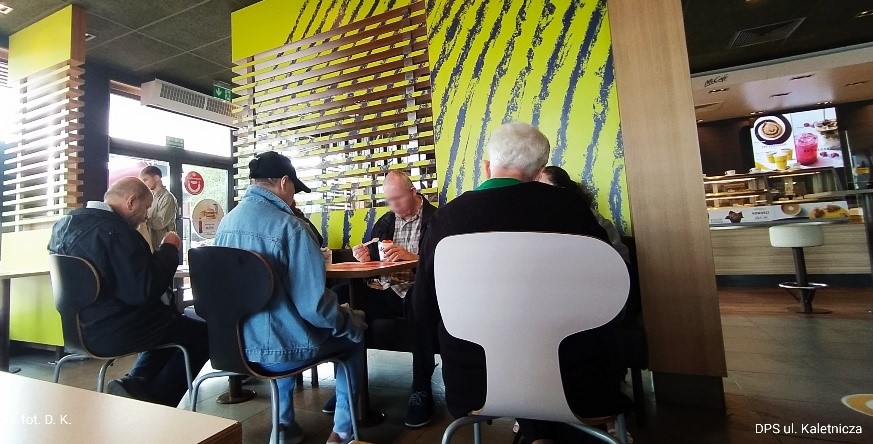 Na zdjęciu sześcioosobowa grupa ludzi siedząca na krzesełkach przy stolikach, jedząca lody w kawiarni