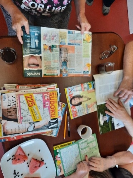 Zdjęcie nr.18 Przedstawia mieszkańców czytających oraz rozwiązujących krzyżówki. Artykuły leżą na brązowym stole.