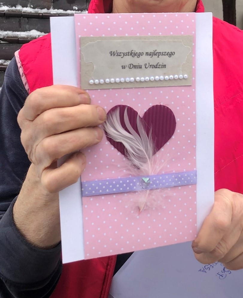 Fotografia przedstawia kartkę urodzinową  w rękach jubilatki. Kartka w różowym kolorze z czerwonym sercem na środku
