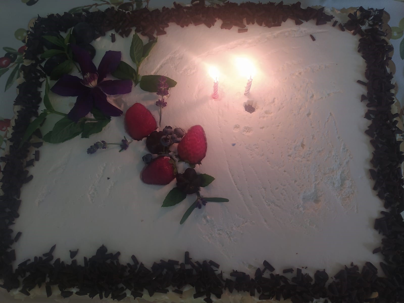 śmietankowy tort urodzinowy udekorowany czerwonymi malinami i wiórkami czekolady. Na torcie palą się 2 świeczki.