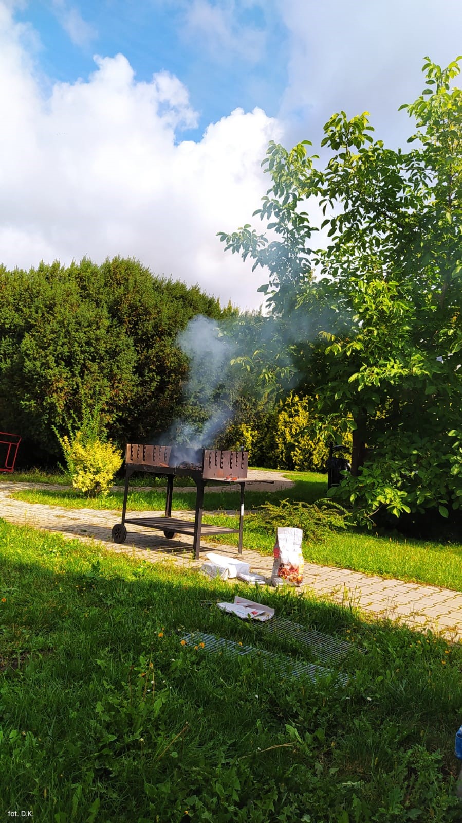 Na zdjęciu zielony ogród z brukowaną ścieżką na której stoi grill z którego unosi się dym. Po prawej stronie grilla papierowa torba z węglem i przybory do rozpalania