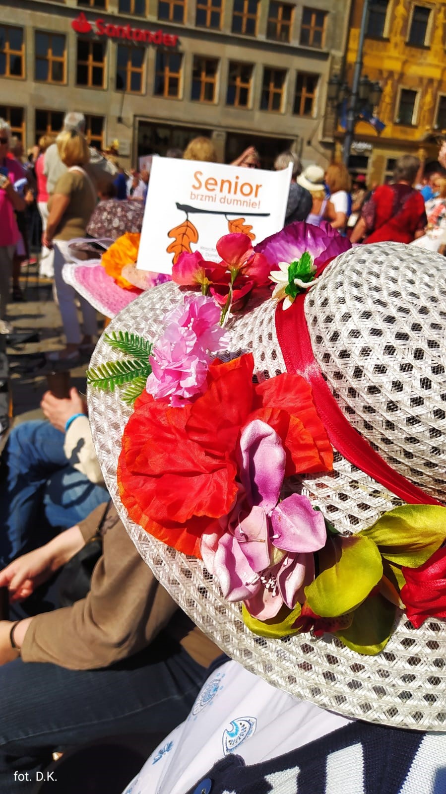 Na zdjęciu, na pierwszym planie beżowy kapelusz ustrojony w kwiaty koloru czerwonego i różowego dookoła tłum ludzi. W centrum chorągiewka z napisem „senior brzmi dumnie”