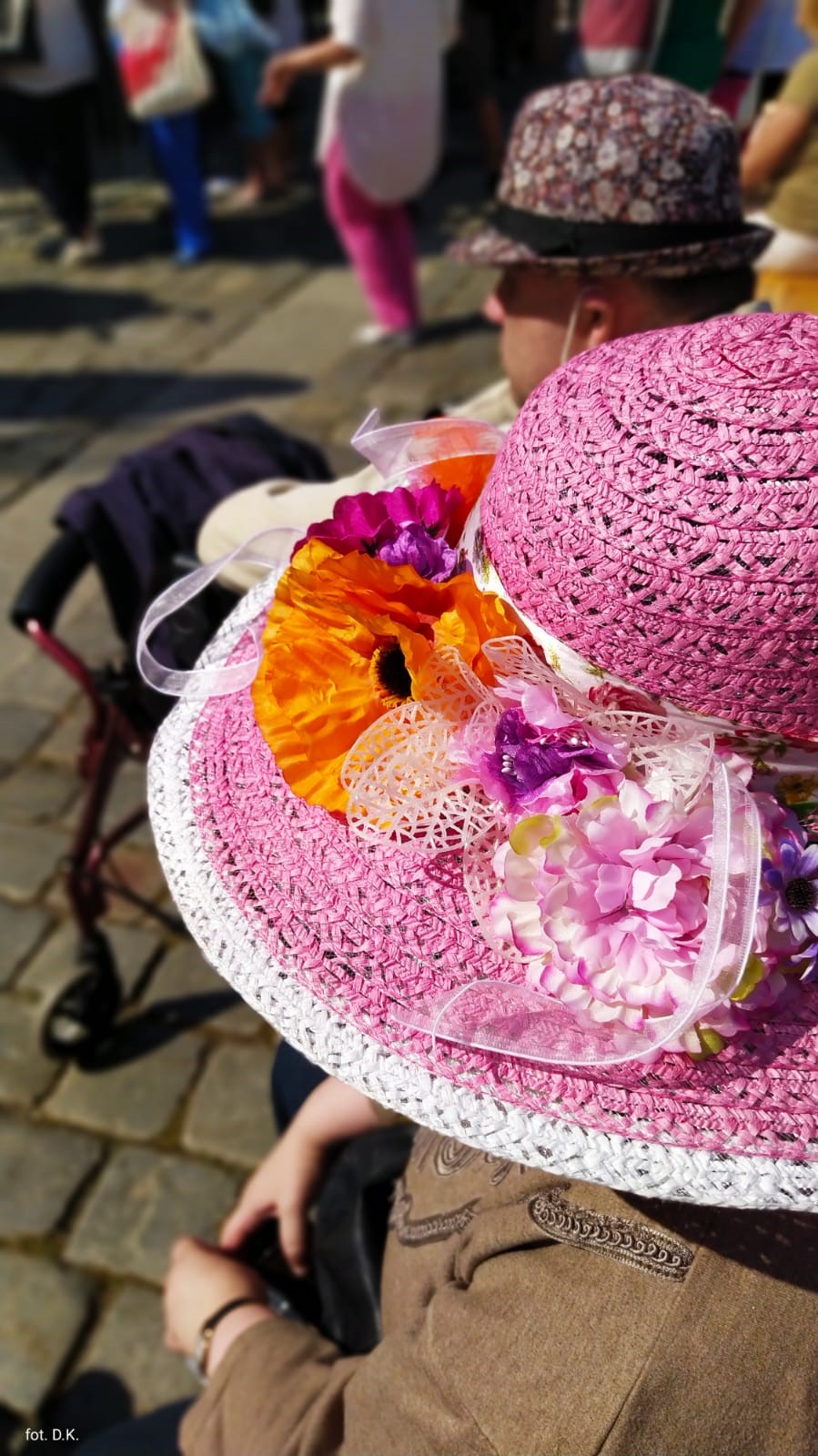 Na zdjęciu w centralnym punkcie różowy kapelusz z kolorowymi kwiatami. W tle rozmyte postacie