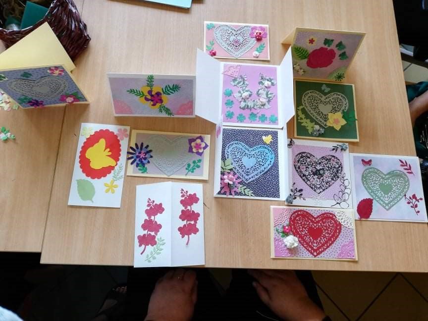Na zdjęciu drewniany stół z ręcznie robionymi kartkami. Kartki leżą w czterech rzędach łącznie leży 13 kartek z motywami serc, kwiatów i motyli