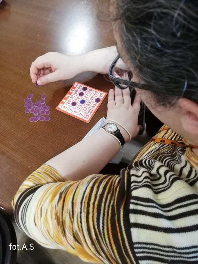 Na zdjęciu ciemny drewniany stół z prawej strony kobieta w brązowo beżowej bluzce przed nią na blacie karta do gry w bingo