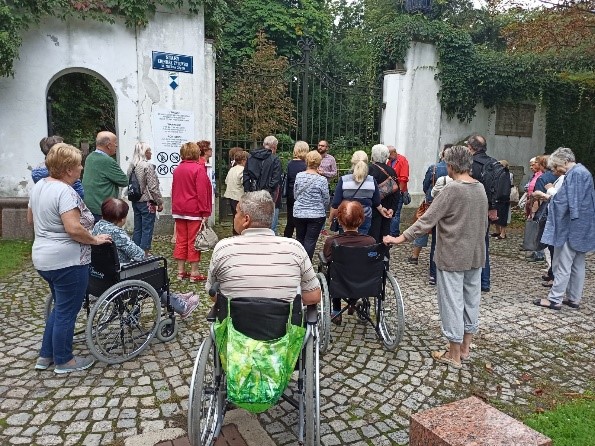 Zdjęcie przedstawia grupę mieszkańców stojących przed brama Cmentarza Żydowskiego. W tle widać białe mury otaczające Cmentarz 