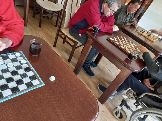 Zdjęcie przedstawia mieszkańców, stoliki a na nich warcaby i szachy